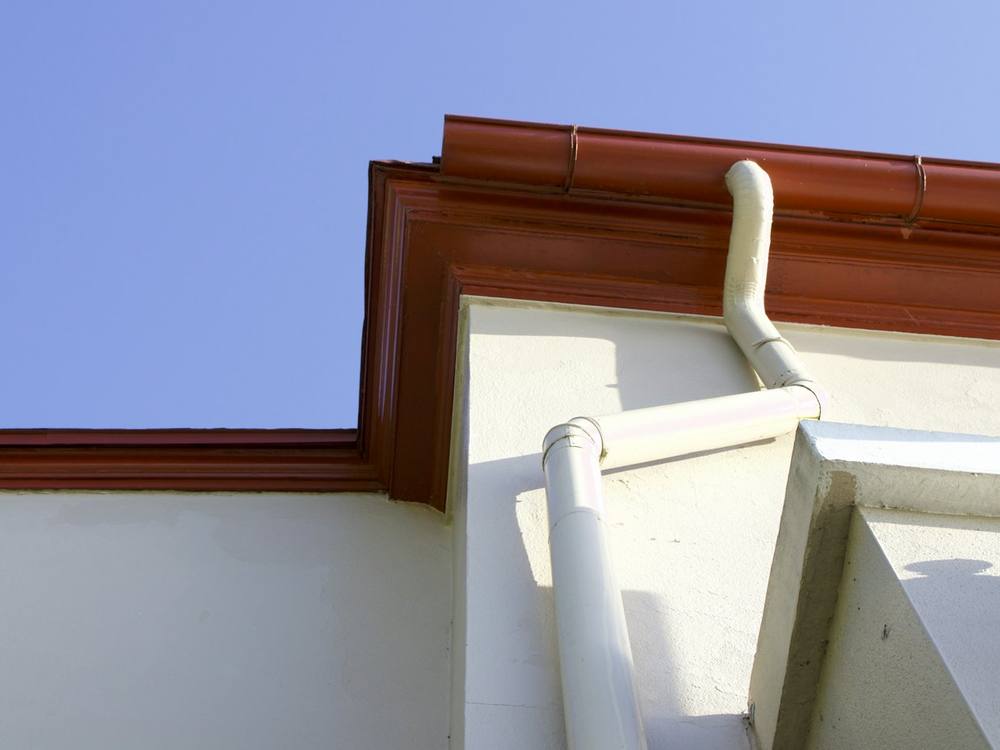 A coloured metal gutter along the eaves of a white rendered house with a matching downpipe